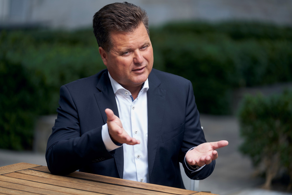 Ein Mann mit kurzen, braunen Haaren, einem blauen Sakko und einem weißen Hemd sitzt an einem Tisch und hält beide Hände geöffnet nach oben.