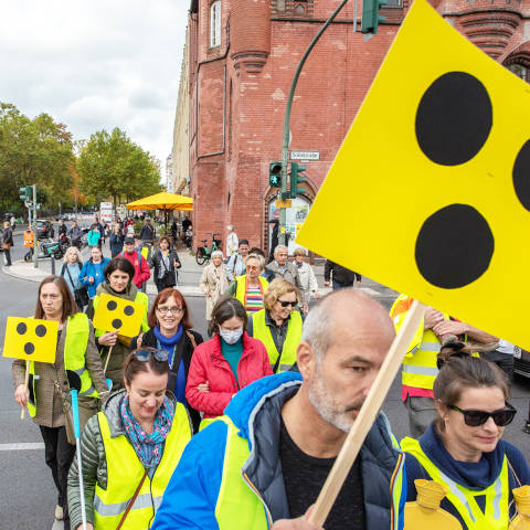 Eine Gruppe von Menschen mit Langstöcken und gelben Plakaten mit drei schwarzen Punkten gehen über die Ampelkreuzung Schloßstraße/Grunewaldstraße.