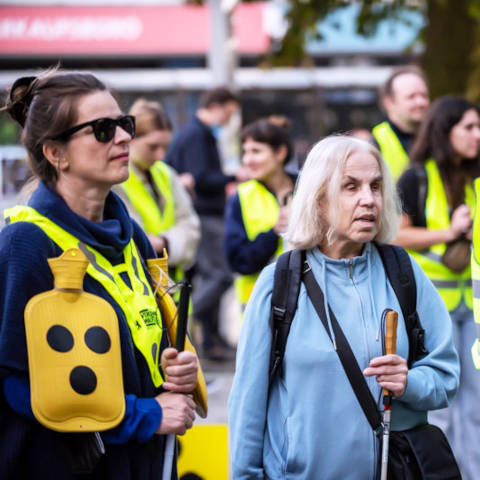 Eine ältere Dame im hellblauen Pullover und eine Jüngere, die mit gelber
Warnweste und gelb bemalter Wärmflasche. Beide halten jeweils einen Langstock in
ihren Händen