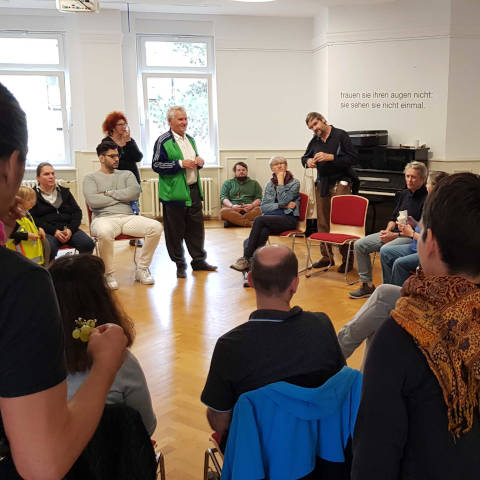 Eine größere Gruppe sitzt und steht im Kreis, darunter die Initiatoren Olaf
Garbow und Gerd Conradt. Über einem Klavier ist an der Wand der Text in großen
Buchstaben angebracht: „Trauen Sie Ihren Augen nicht – Sie sehen sie nicht einmal“