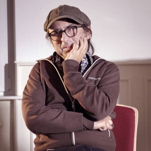Frank Amann trägt eine braune Schirmmütze, eine Brille und eine braune
Kapuzenjacke. Er schaut nach links und stützt sein Kinn in seine Hand.