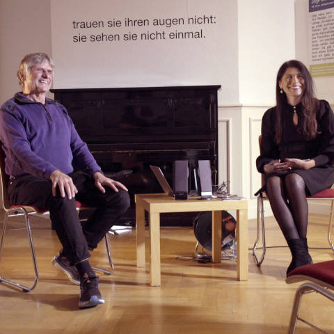 Eberhard Dietrich hat mittellange weiße Haare, trägt einen blauen Pullover
und lächelt. Rechts daneben sitzt Llane Fragoso Maldonaldo, sie hat schwarze lange
Haare, trägt ein schwarzes Kleid und lächelt. Hinter ihnen steht auf der Wand „Trauen
sie ihren Augen nicht, sie können sie nicht einmal sehen.