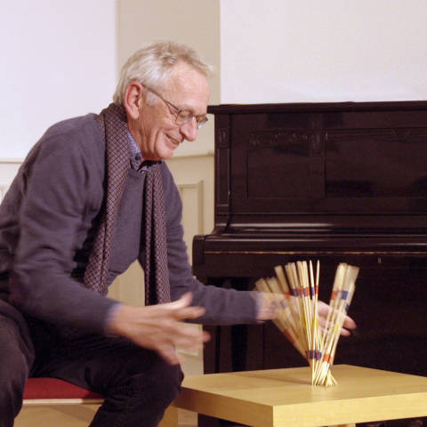 Prof. Dr. Peter Bexte hat kurze weiße Haare, trägt eine Brille. Aus seiner
Hand fallen Mikadostäbchen auf den Tisch.