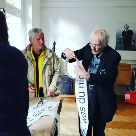 Gert Conradt und Timm Ulrichs stehen im Raum und arbeiten an einem Schriftzug
mit Klebe Buchstaben, ersichtlich ist der Text: „Was du nicht….“
